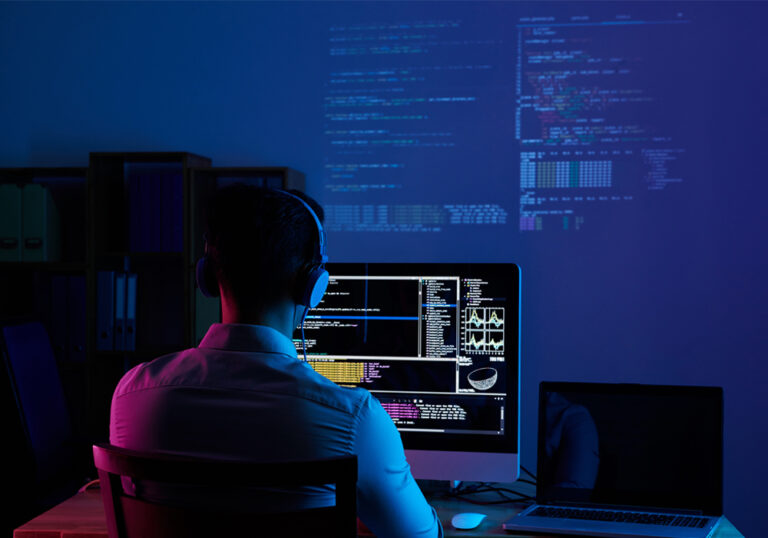 Developer wearing headphones coding at night with data and code displayed on a large monitor and projected on the wall